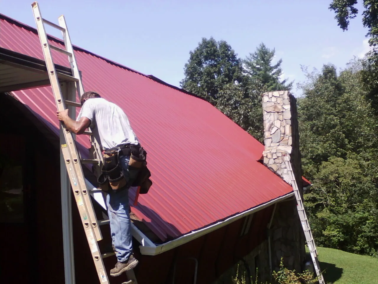 Roofer on a ladder beside a red metal roof and stone chimney — Soffit & Fascia Repair by Roofing Brothers in Ellenboro